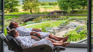 A couple relaxing on wicker lounge chairs by a large window overlooking a landscaped garden and outdoor seating area. - Lodore Falls Hotel (Keswick)