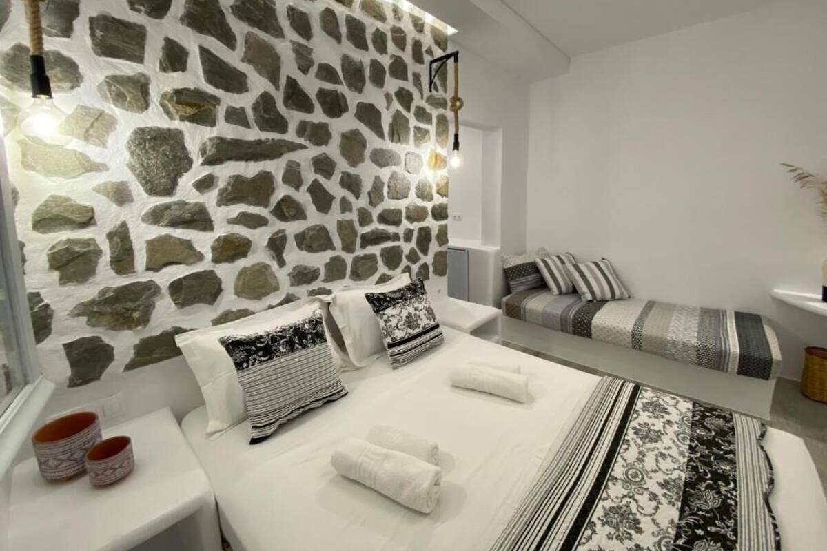 Ein modernes Schlafzimmer mit einem Kopfteil aus Stein, einem Bett mit weißen Bettwäsche, einem Nachttisch mit Lampe und einem Sofa mit gemusterten Kissen.