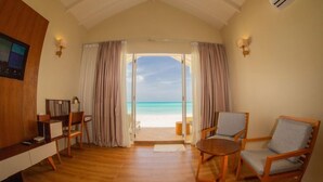 Una habitación con suelo de madera, un escritorio con una silla, dos sillones y vista a la playa a través de una puerta.