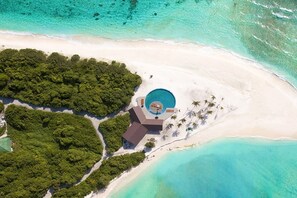 Ovenudblik over en feriebolig med en central pool, omgivet af frodig vegetation og en sandstrand.