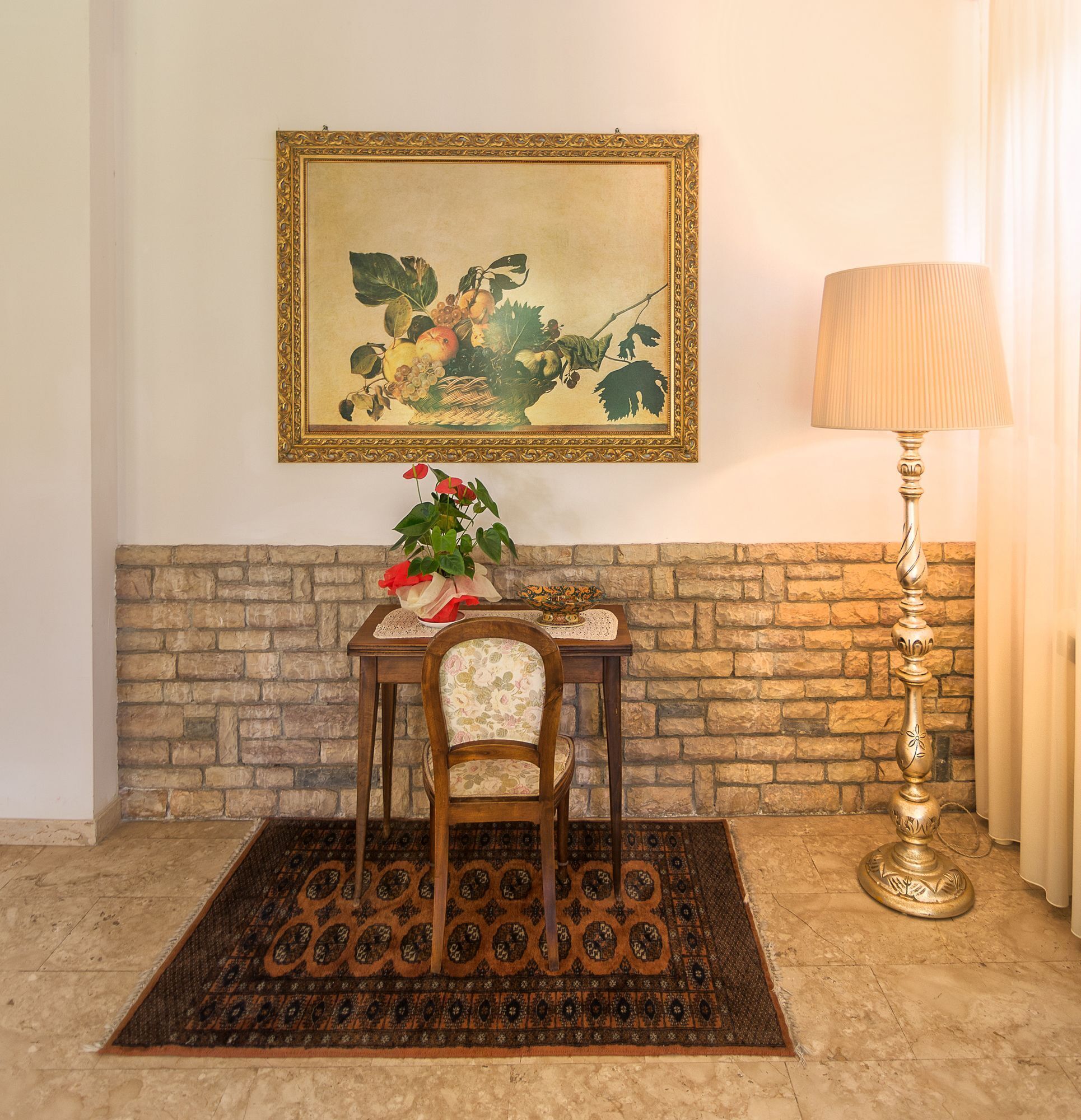 A room with a stone wall, a framed painting, a table with a vase of flowers, a chair, a patterned rug, and a lamp.