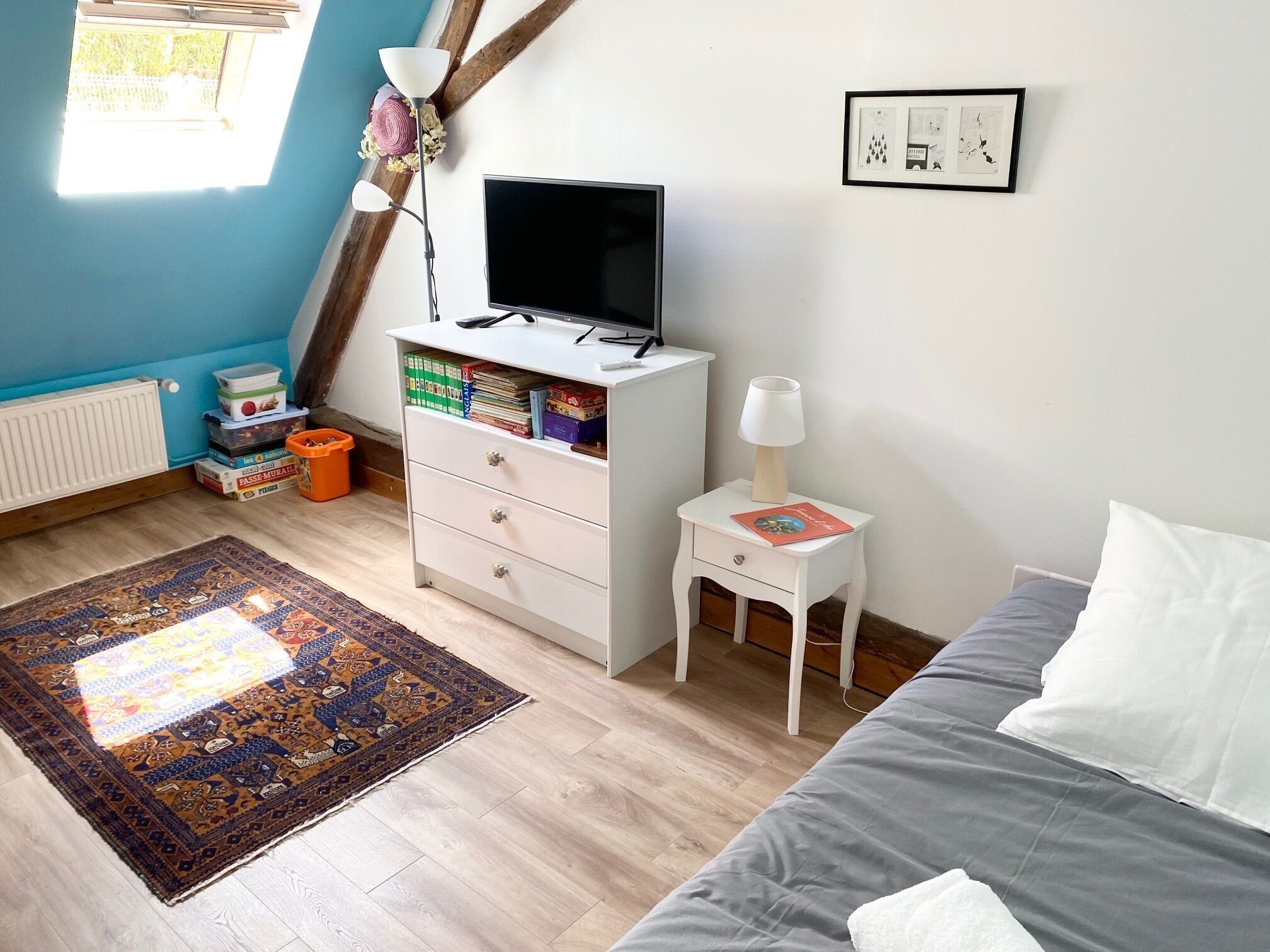 Een slaapkamer met een bed, een bureau met een computer, een boekenplank, een raam met zonwering en een radiator.