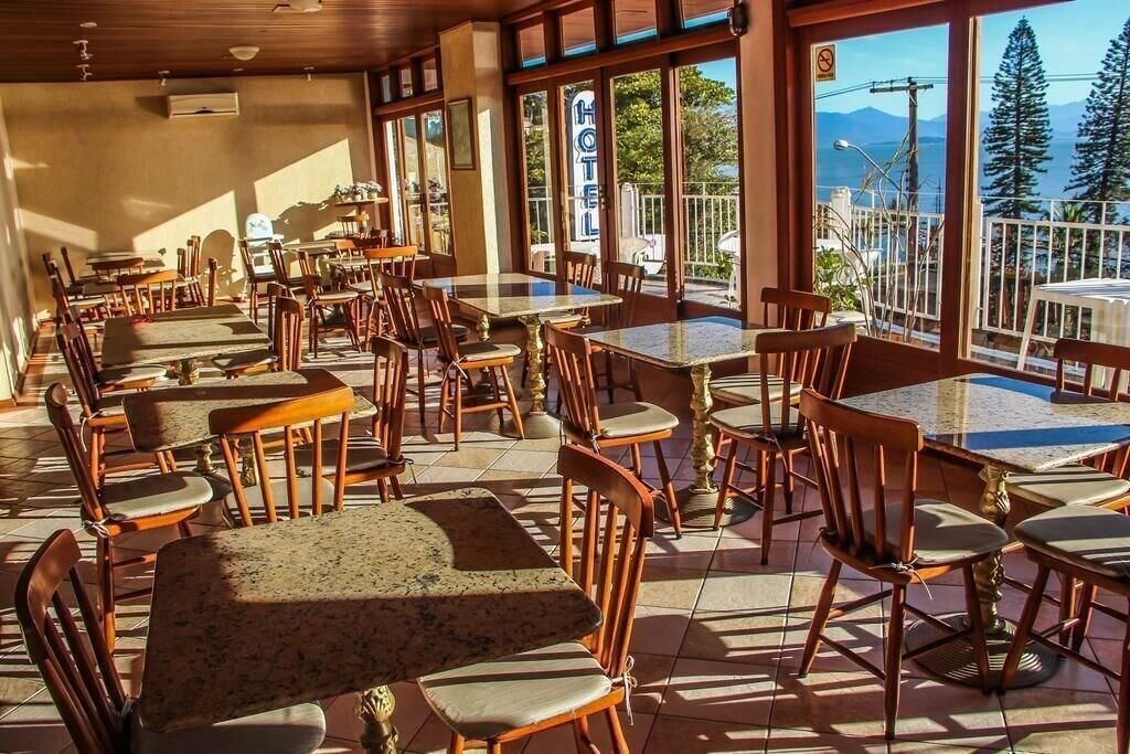 A restaurant with outdoor seating, wooden tables and chairs, and large windows offering a view of the sea and mountains.