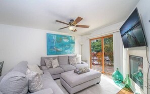 A living room with a grey sectional sofa, a ceiling fan, a large window with a view of greenery, and a flat-screen TV mounted on the wall.