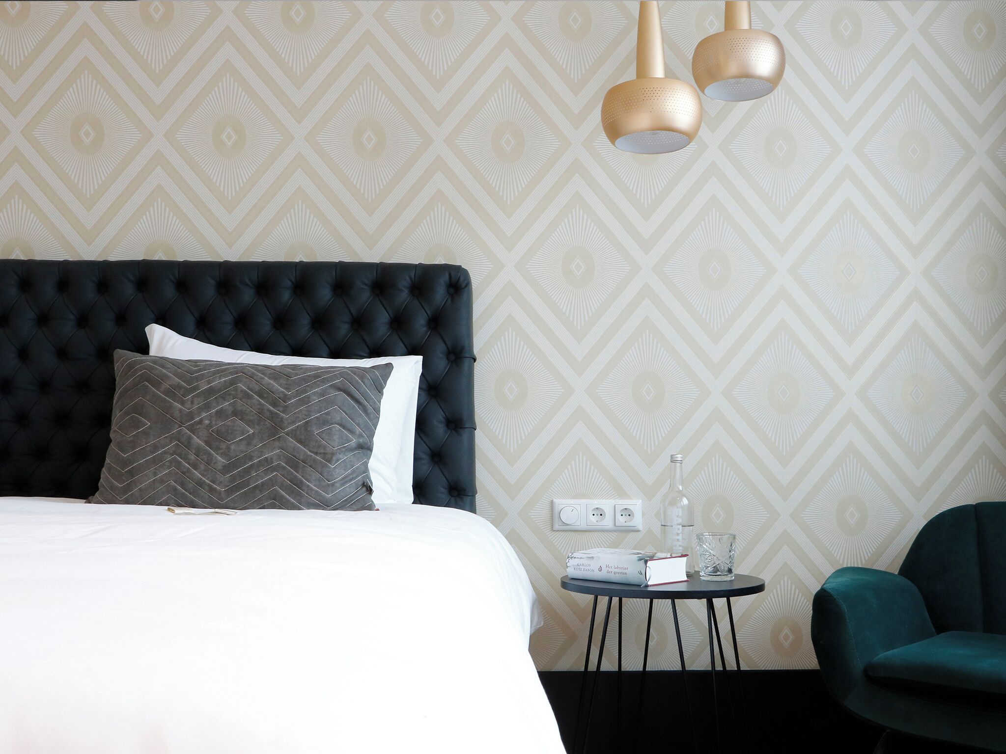 A hotel room with a tufted headboard, a bed with white and gray linens, a small round table with a book and glass, and a teal armchair.