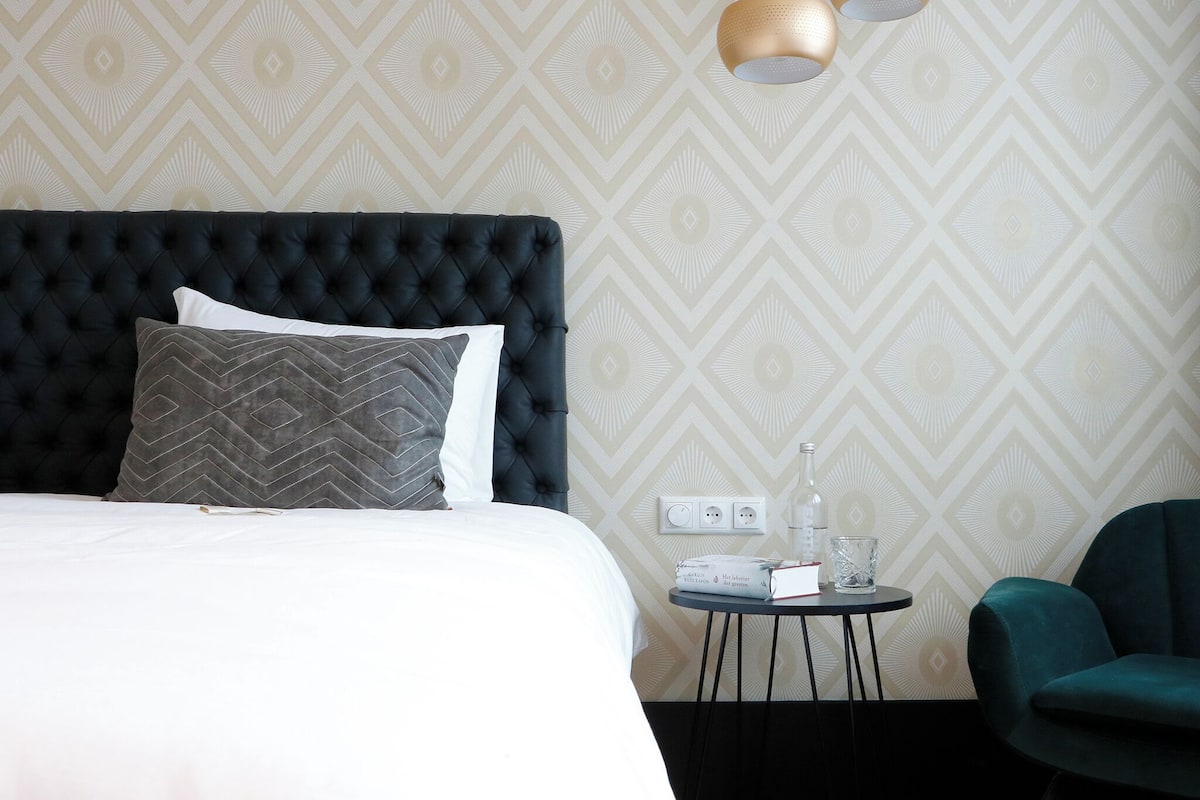 A hotel room with a tufted headboard, a bed with white and gray linens, a small round table with a book and glass, and a teal armchair.