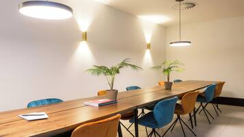 A conference room with a long wooden table, blue and orange chairs, two potted plants, and pendant lighting.