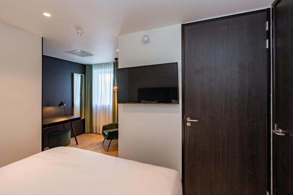 A hotel room with a bed, a desk with a chair, a television mounted on the wall, and a dark wooden wardrobe.