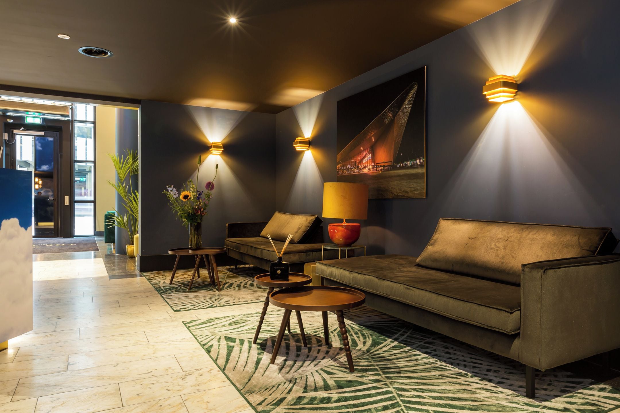 Een moderne lobby met een bank, twee bijzettafels, een lamp en wandlampen.