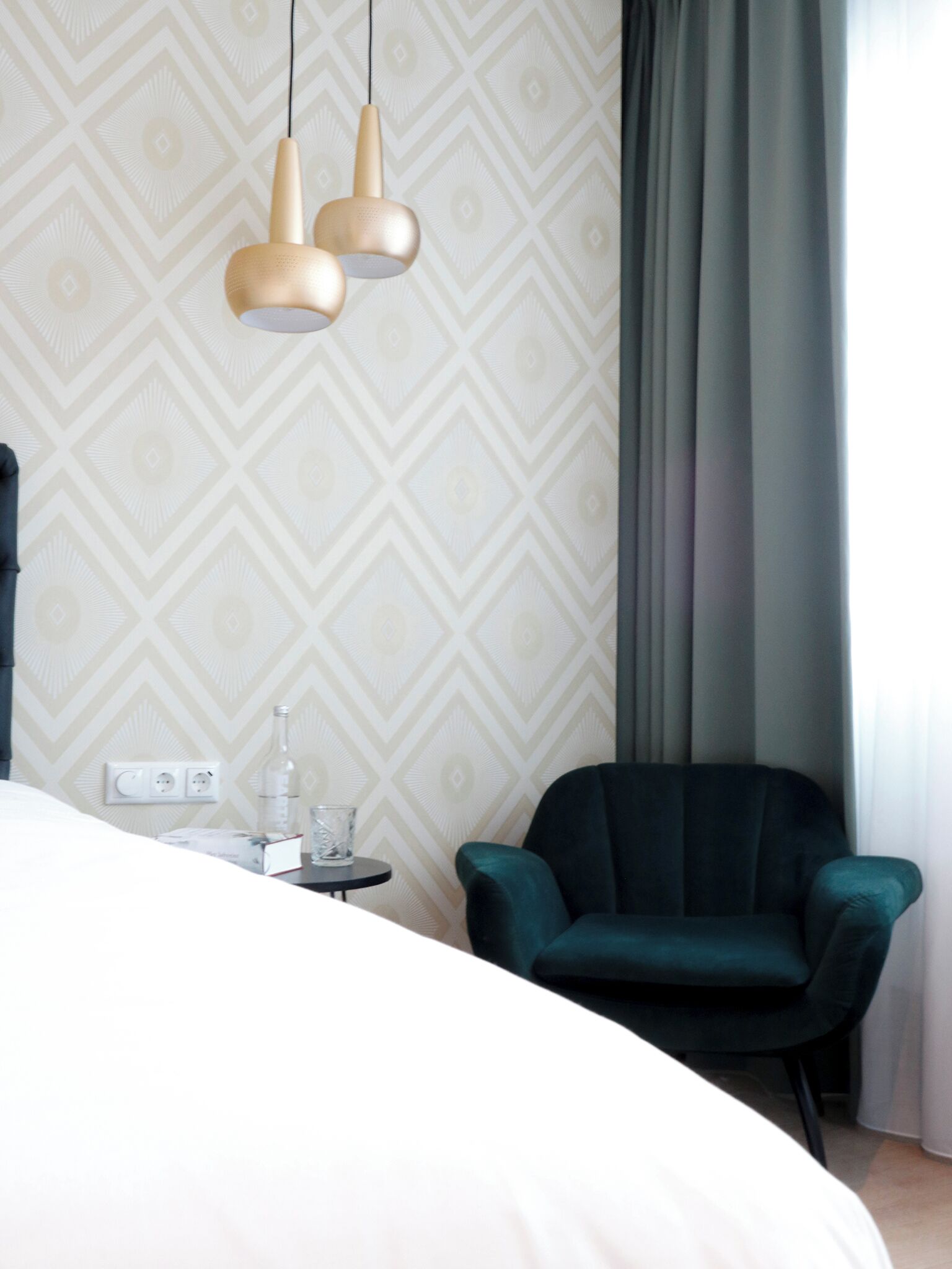 A hotel room with a bed, a chair, a small table with a bottle and glass, and a wall with a geometric pattern.