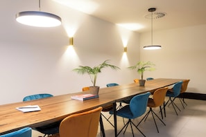 A conference room with a long wooden table, blue and orange chairs, two potted plants, and pendant lighting. - The James Hotel Rotterdam (Rotterdam)
