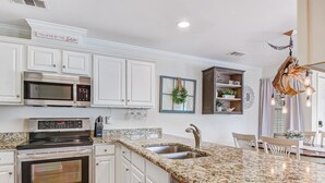 A modern kitchen with white cabinetry, stainless steel appliances, a granite countertop, and a dining area with a chandelier. - Heated Pool Access,Walk to Beach (Tybee Island)