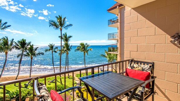 A balcony with a table and chairs, overlooking a beach with palm trees and the ocean. - Kealia 402 Oceanfront Condo and Beautiful Secluded White-Sand Sugar Beach (Kihei)