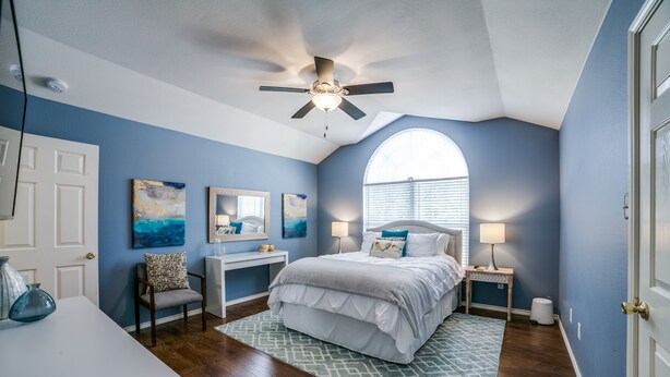 A bedroom with a bed, a desk, a chair, a ceiling fan, and a window with blinds.