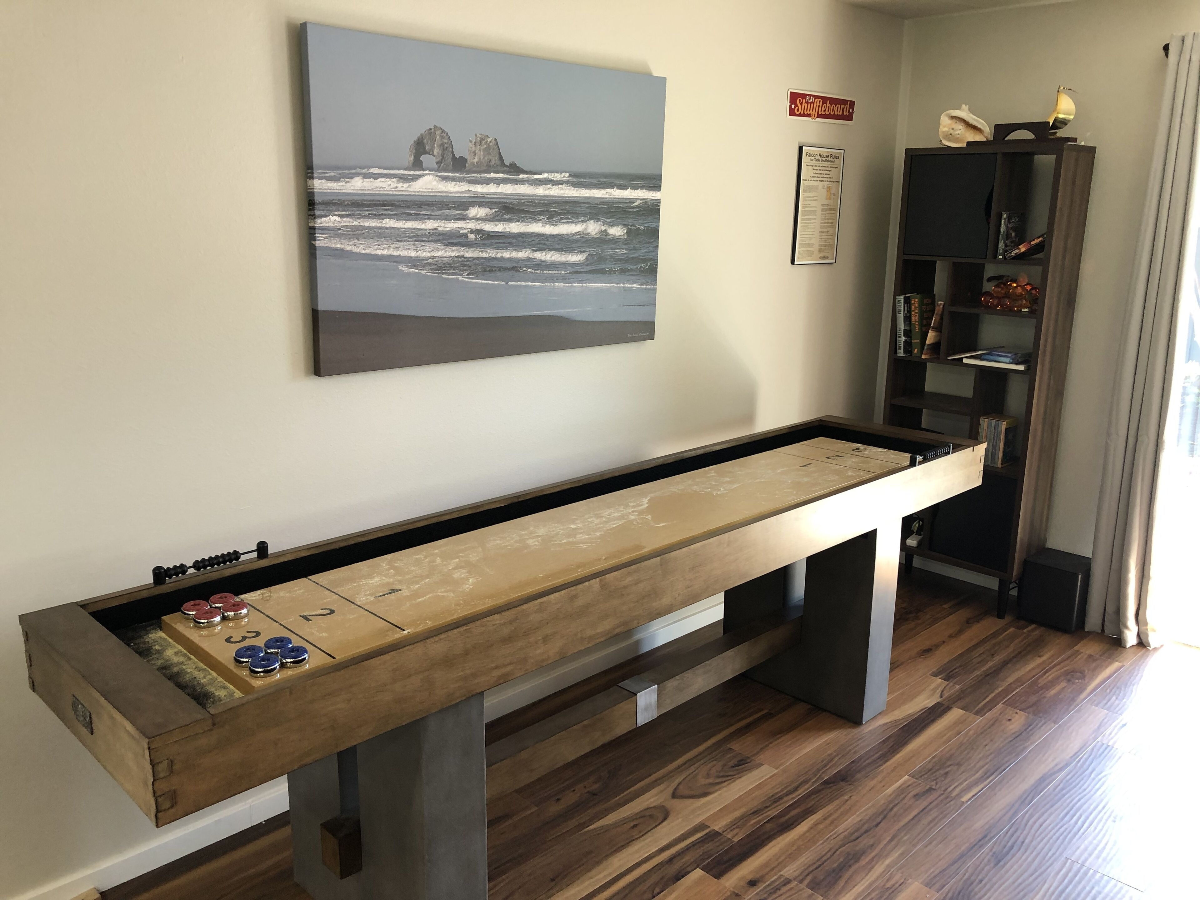 Une table de jeu de shuffleboard dans une pièce dotée d’un plancher en bois, un tableau représentant un paysage côtier et une bibliothèque contenant divers objets.