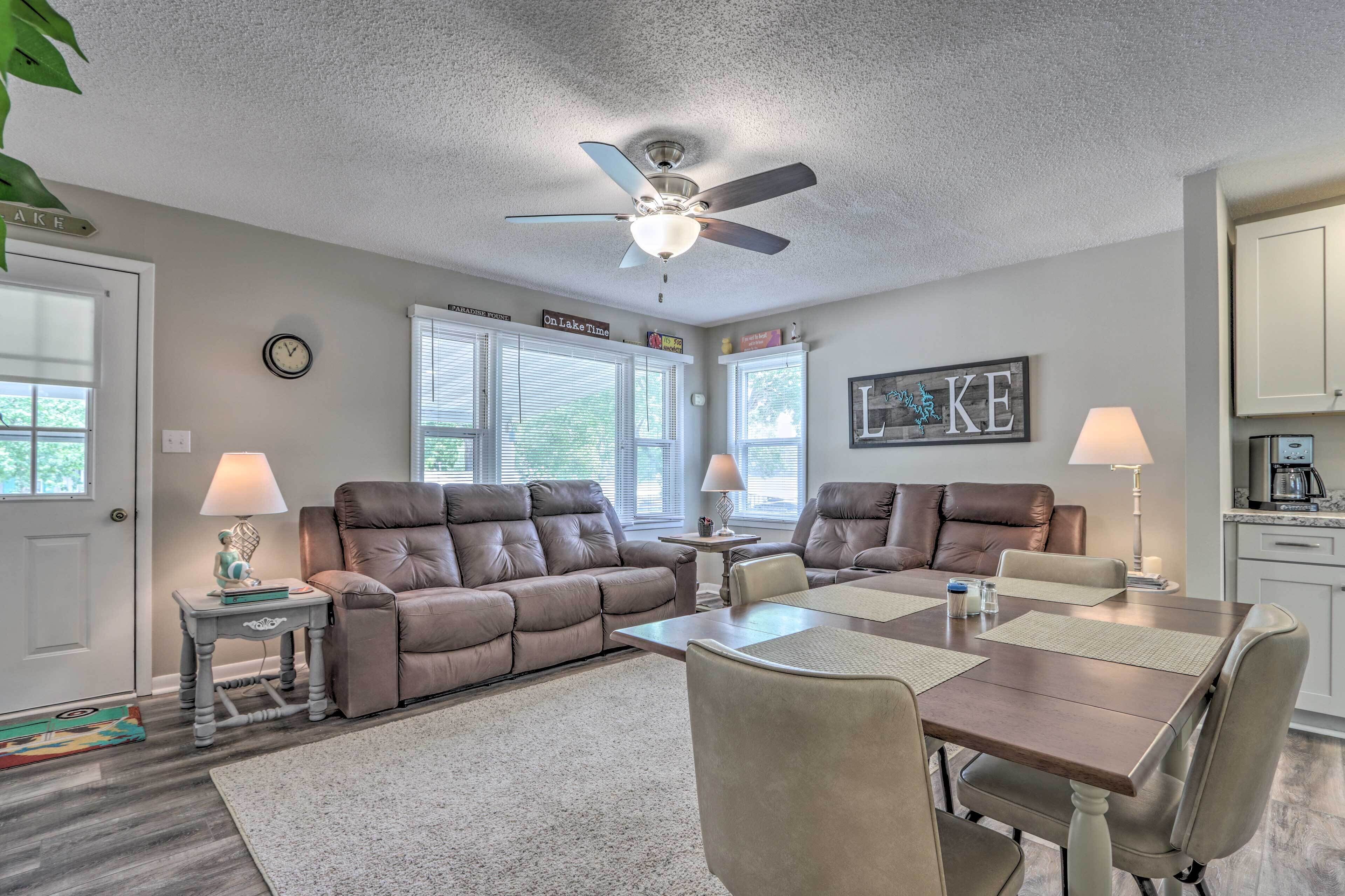A spacious living room with a brown leather sofa, a dining table with four chairs, a ceiling fan, and a kitchen area with cabinets and a coffee maker.