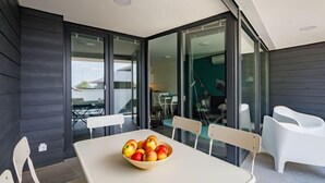 Un espace repas moderne avec une table et des chaises blanches, un saladier de pommes et une vue sur une terrasse extérieure, accessible par de grandes portes vitrées.