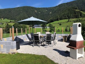 Eine Terrass mit Steintisch und -stühlen, einem Grill und einem Sonnenschirm.
