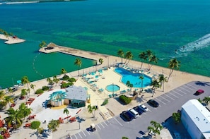 An aerial view of a resort with a large swimming pool, palm trees, and a parking area.