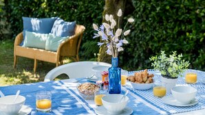 Ein Tisch im Freien mit Frühstücksangeboten, darunter Croissants, Müsli und Saft, sowie eine blaue Vase mit Blumen.