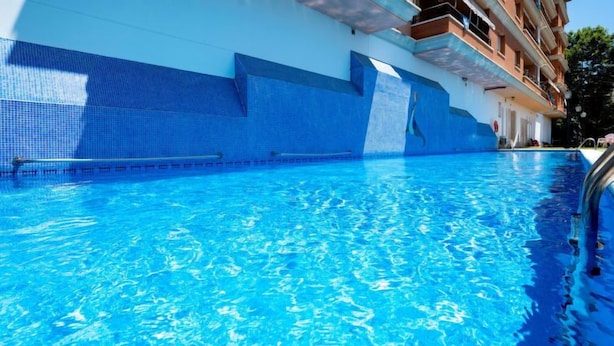 A swimming pool with clear blue water, a tiled edge, and a building with balconies in the background.