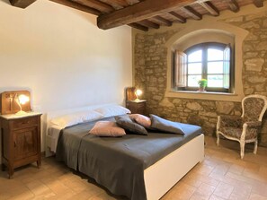 Quarto com parede de pedra, janela com vista, cama com almofadas, cadeira e abajures.
