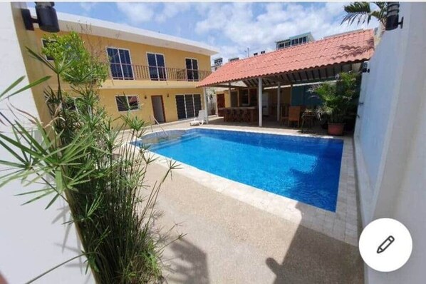 A swimming pool with a tiled deck, surrounded by a building with a red-tiled roof and a yellow facade.