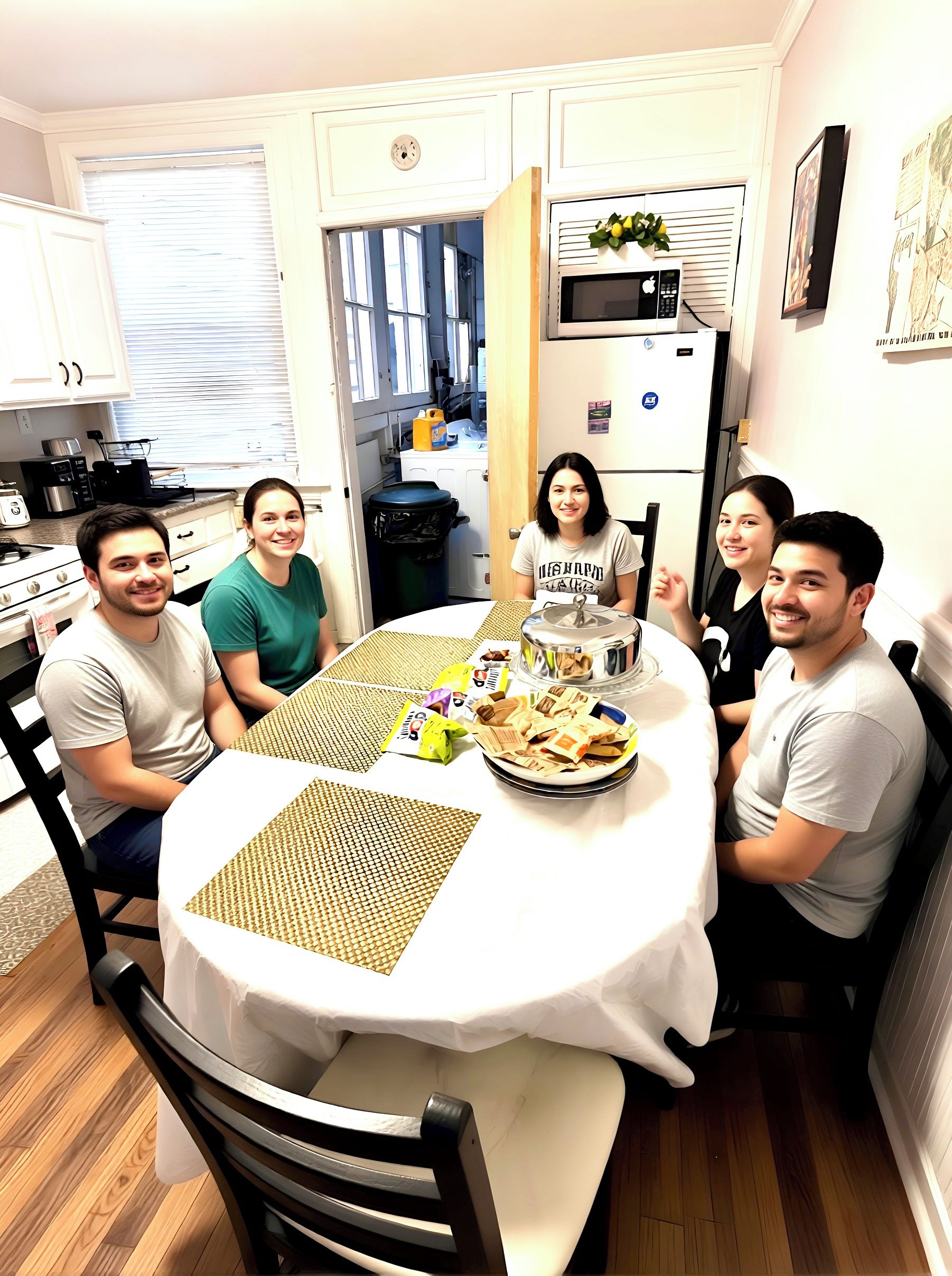 A group of people seated around a round table with a white tablecloth, in a kitchen with white cabinets and a microwave.