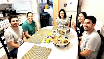 A group of people seated around a round table with a white tablecloth, in a kitchen with white cabinets and a microwave.
