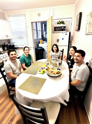 A group of people seated around a round table with a white tablecloth, in a kitchen with white cabinets and a microwave.