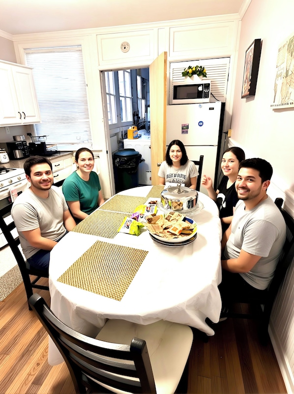 A group of people seated around a round table with a white tablecloth, in a kitchen with white cabinets and a microwave.