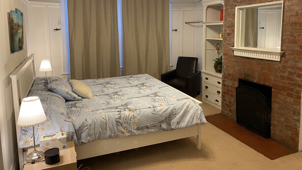 Une chambre à coucher comprenant un lit, une table de chevet, une lampe, une chaise, une cheminée et un miroir.