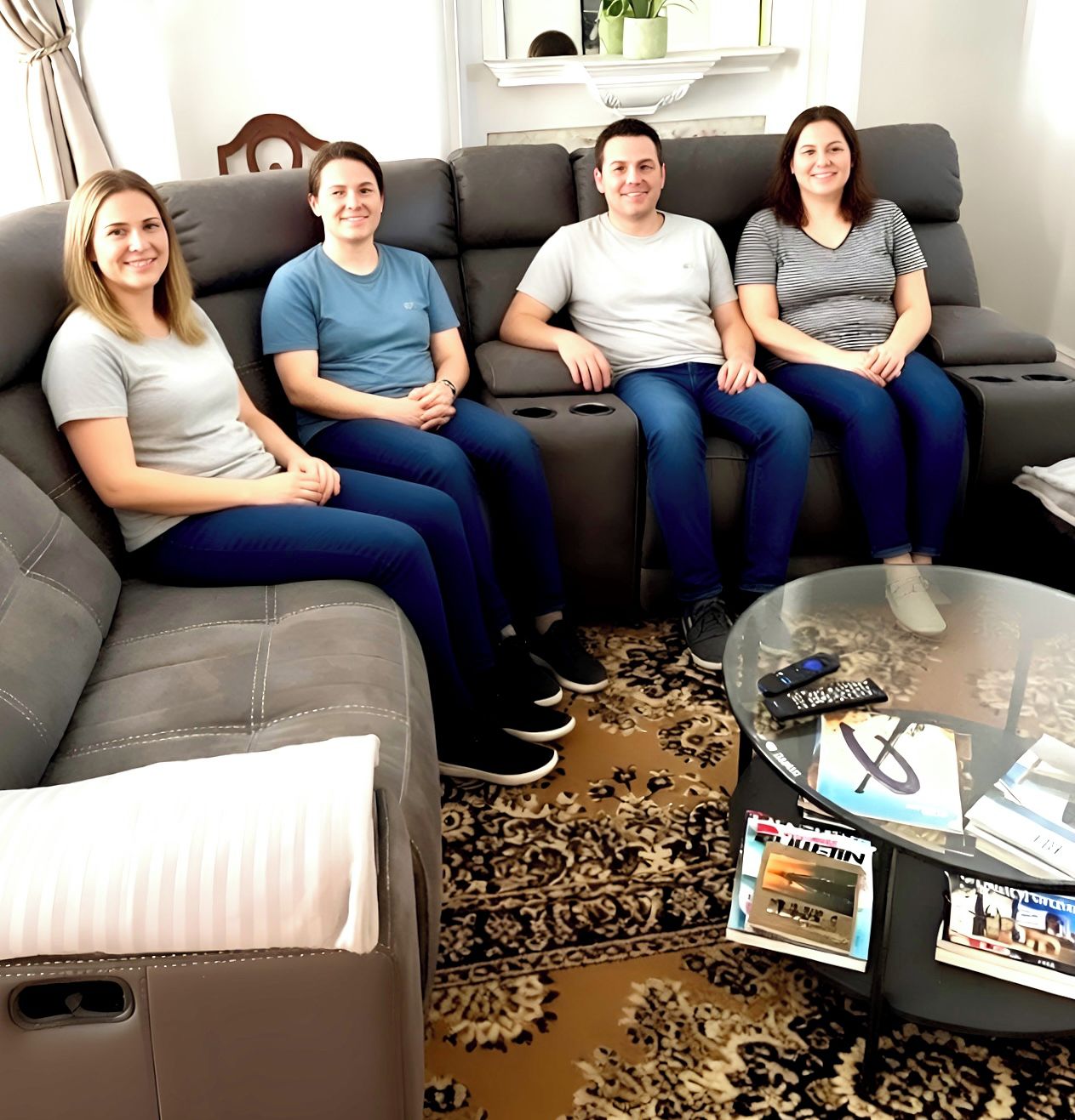 A living room with a grey sofa, a glass coffee table with magazines, and a patterned rug.