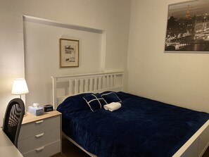Ein Schlafzimmer mit einem Bett, einem Nachttisch, einer Lampe, einem Sessel, einem gerahmten Bild und einem Fenster mit Blick auf eine Stadtlandschaft.
