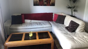 A living room with a beige sectional sofa, a glass-top coffee table, a red throw blanket, and a painting of two figures on the wall. - Immaculate 3-Bed Apartment in Kilkenny (Kilkenny)