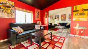 A modern living room with a black leather sofa, a coffee table, and red and white patterned rugs. There are movie posters on the walls and a small table with a wine glass and a bottle. - Stylish Desert Retreat - Stunning Views, 3 Min to Joshua Tree Park (Twentynine Palms)