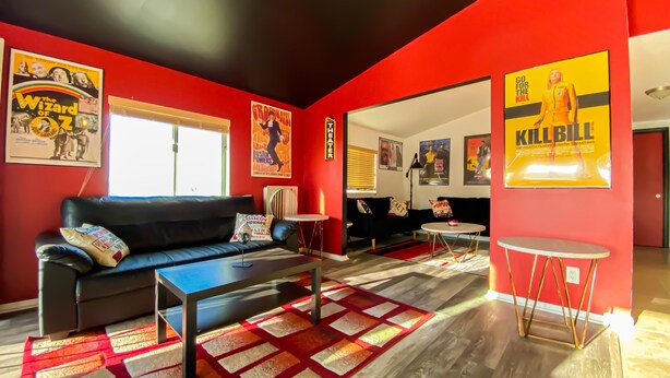 A modern living room with red walls, a black leather sofa, a coffee table, and a small round table. There are posters on the walls and a window with blinds. - Stylish Desert Retreat - Stunning Views, 3 Min to Joshua Tree Park (Twentynine Palms)
