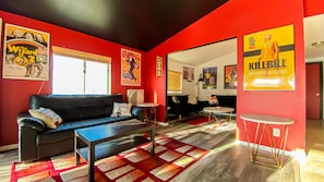 A modern living room with red walls, a black leather sofa, a coffee table, and a small round table. There are posters on the walls and a window with blinds.