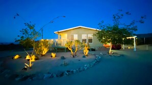 A house with a porch, a covered patio, and a landscaped garden with rocks and small trees. - Stylish Desert Retreat - Stunning Views, 3 Min to Joshua Tree Park (Twentynine Palms)