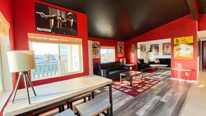 A modern living room with red walls, a black leather sofa, a wooden coffee table, and a dining table with benches. There is a large window providing a view of the outdoors. - Stylish Desert Retreat - Stunning Views, 3 Min to Joshua Tree Park (Twentynine Palms)