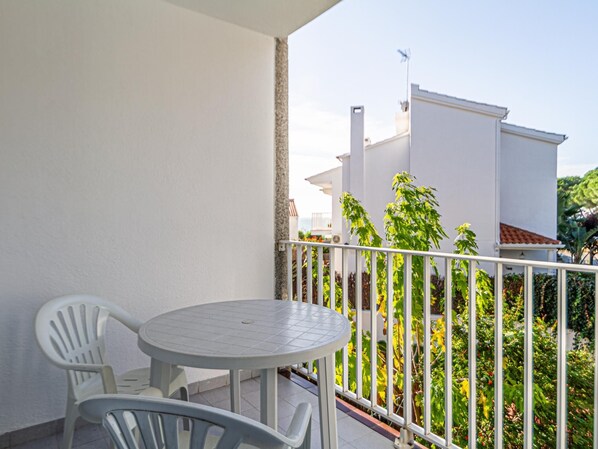 A balcony with a round table and two chairs, a white railing, and a view of a building and greenery.