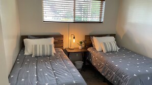 A bedroom with two single beds, a nightstand with a lamp, a small potted plant, and a window with blinds. - Newly Renovated Mother-in-Law Apartment (Orem)