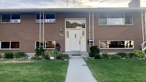 A two-story brick building with a white door and windows, a small garden with grass and plants, and a clear sky. - Newly Renovated Mother-in-Law Apartment (Orem)