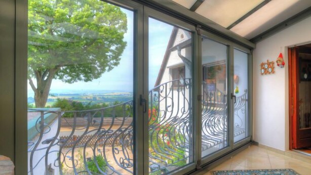 A room with large sliding glass doors leading to a balcony with a metal railing, a view of a tree and landscape, and a patterned rug on the floor. - Am Dohlenberg + Steintisch by Interhome (Schwalenberg)
