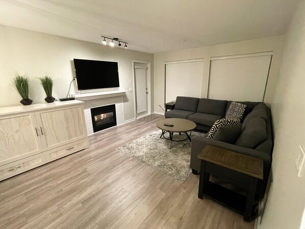 A modern living room with a fireplace, a flat-screen TV mounted above it, a grey sofa, a coffee table, and a cabinet with a plant on top.