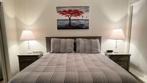 A neatly made bed with a patterned bedspread, two bedside tables with lamps, a wall-mounted painting of a tree, and a nightstand with a clock. - Modern, Deluxe Stoney Creek Northstar Condo. Pool & Hot Tub, Gas Fireplace (Whistler)