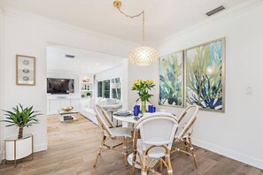 A dining area with wicker chairs, a round table, and a hanging light fixture. There are paintings on the wall and a plant on the side. - The Pearl I By Hotel Home Stays (West Palm Beach)