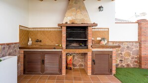An outdoor kitchen with a stone chimney, wooden cabinets, and a tiled backsplash. - Apartment Casa Patricia Nerja with Mountain View, Pool (Nerja)