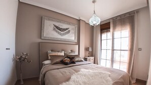 A bedroom with a large bed, a chandelier, a framed artwork above the bed, a nightstand with a lamp, and a window with curtains. - House "Amazing Duplex - 30m from the beach" with Sea View, Terrace, A/C & Wi-Fi (San Juan de los Terreros)