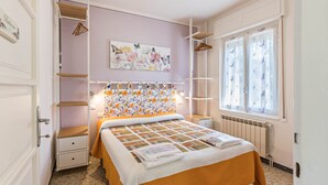A bedroom with a bed, a radiator, a window with curtains, a wall with a floral painting, and a shelf with hanging clothes.